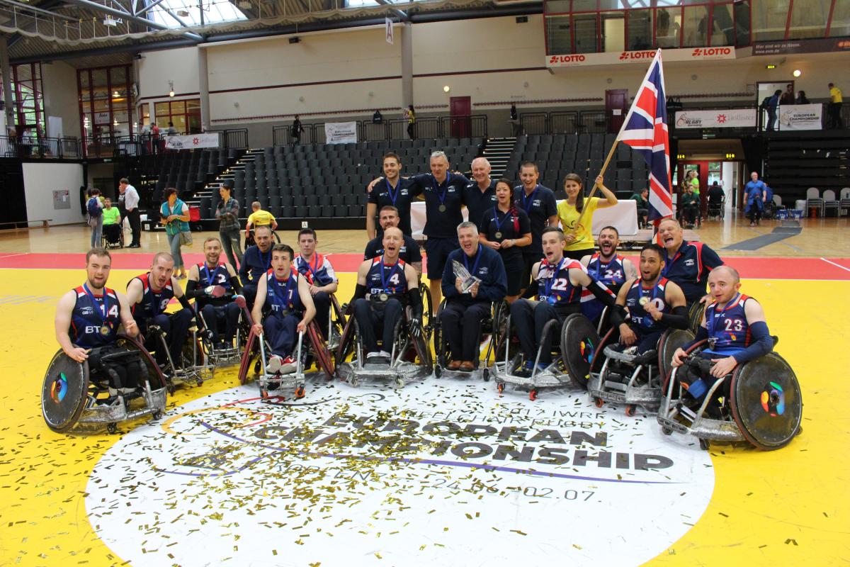 A group of men in wheelchairs surrounded by confetti and ribbons celebrate with the trophy