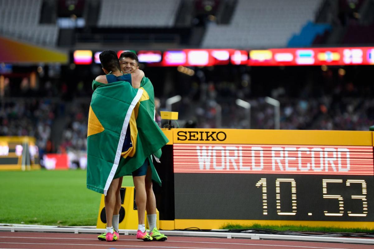 Two athletes hug on the track in front of a bulletin board showing 'WR'