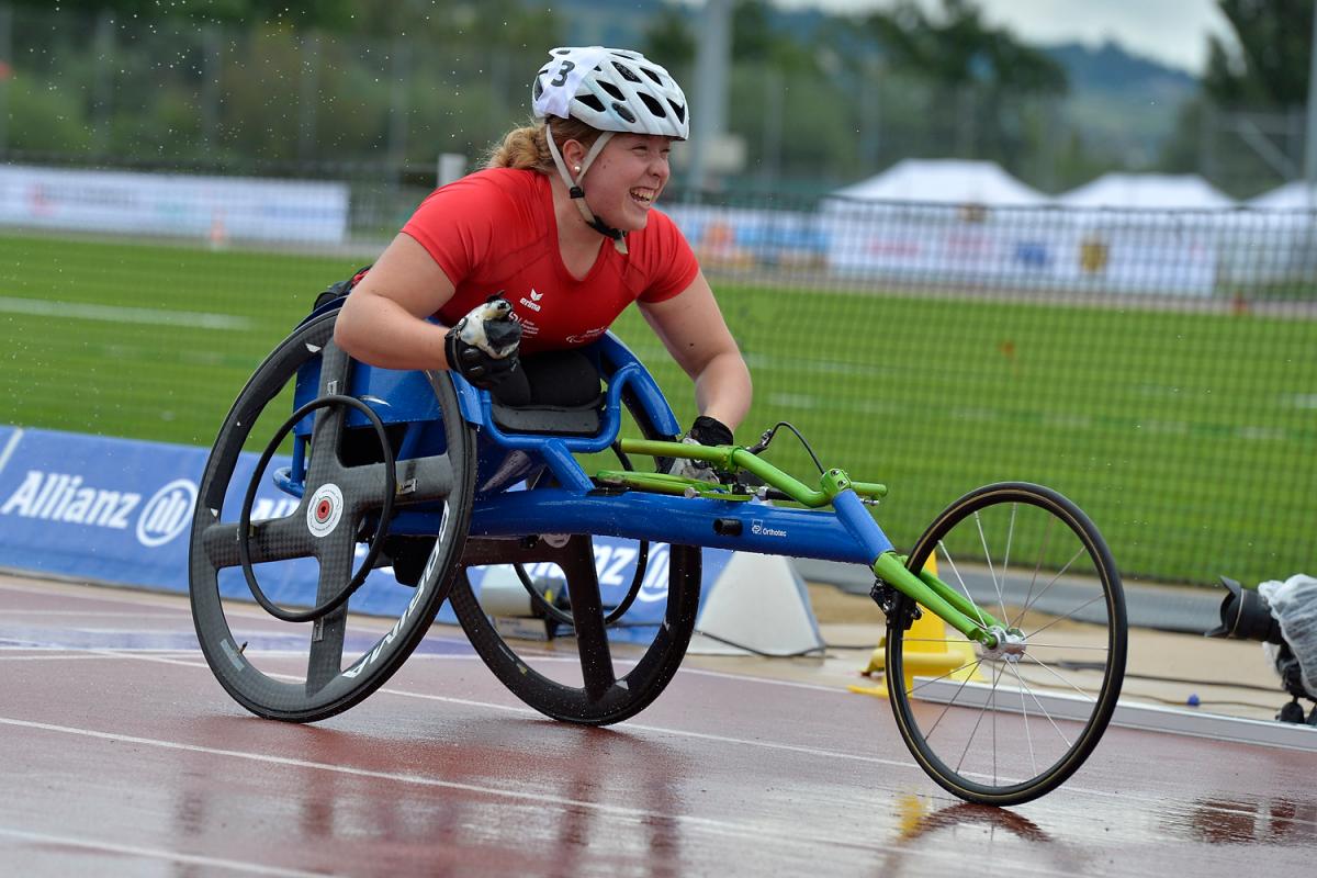 Athlete in a wheelchair celebrates clencing her first with a big smile on a wet track.