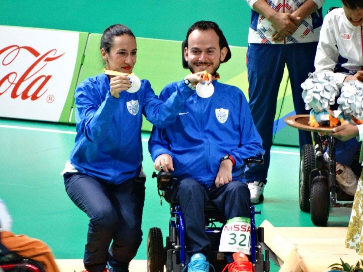 a woman and a man in a wheelchair hold up their silver Paralympic medals