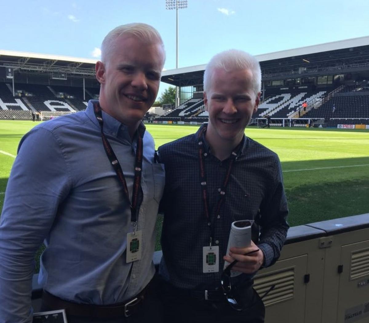 two men smile at the side of a football pitch