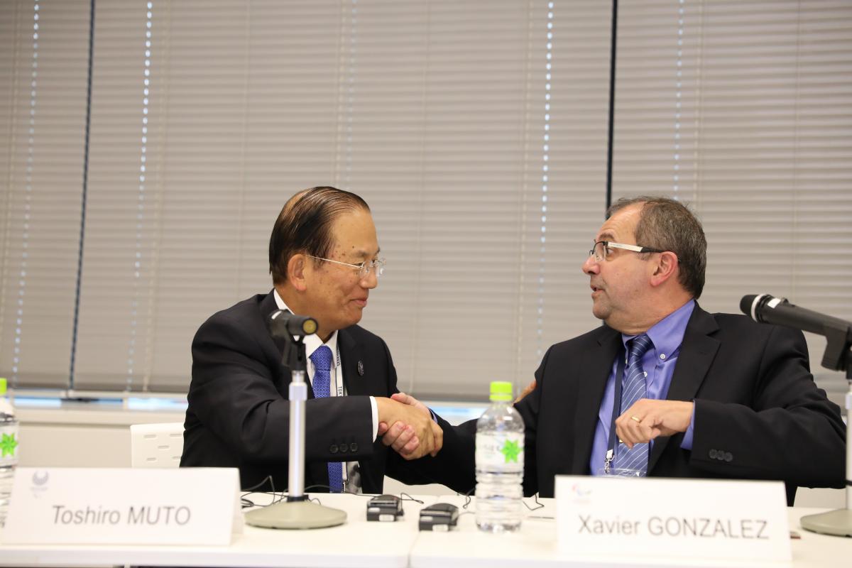 two men shake hands across a table and smile