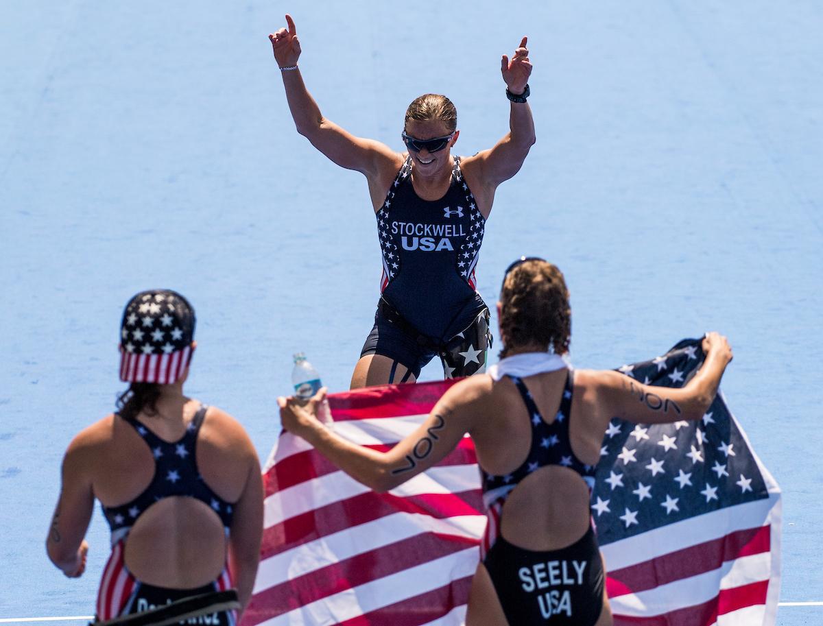 Two women holding the US flag waiting for their teammate to cross the finish line