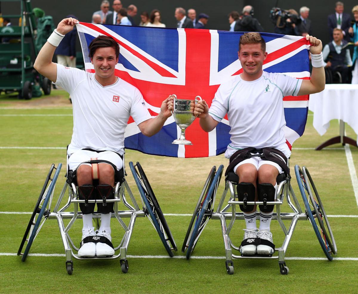 two male wheelchair tennis players hold their trophy and the British flag