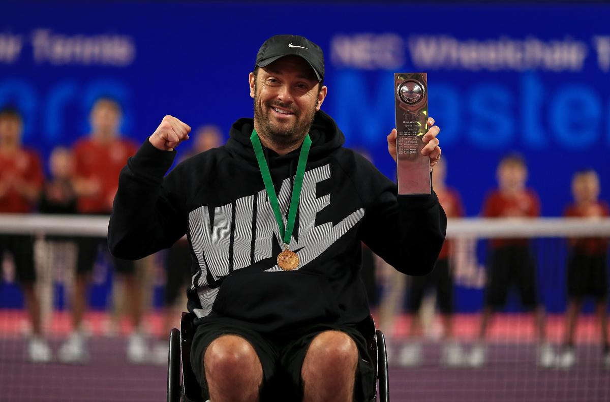 a male wheelchair tennis player holds up a trophy