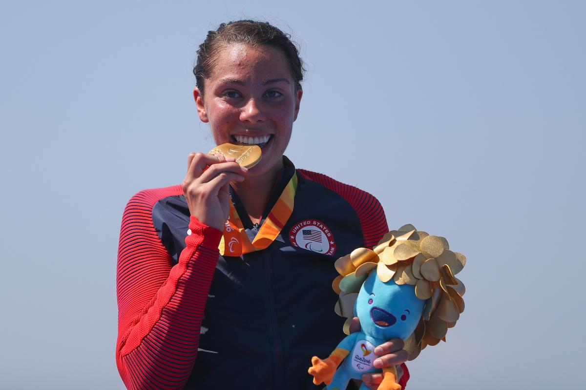 a female Para triathlete stands on the podium and bites her gold medal