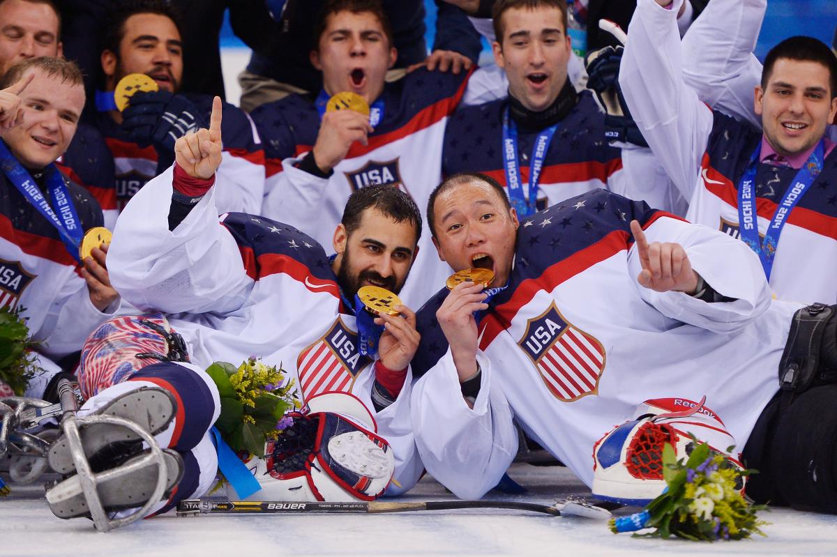 a group of male para ice hockey players lie on the ice and bite their medals in celebration