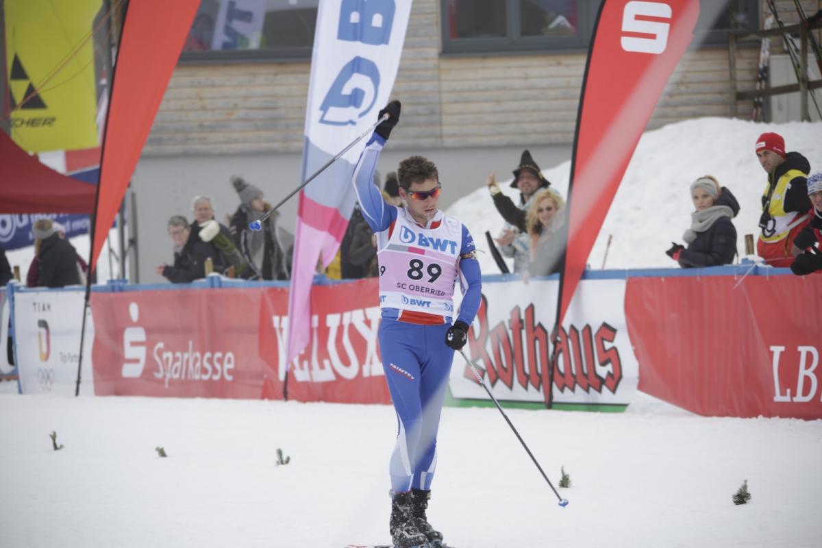 a male Para Nordic skier raises his arms in celebration as he crosses the finish line