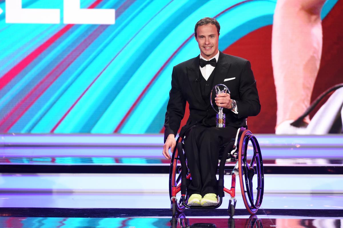Man in wheelchair dressed in a suit holding a trophy