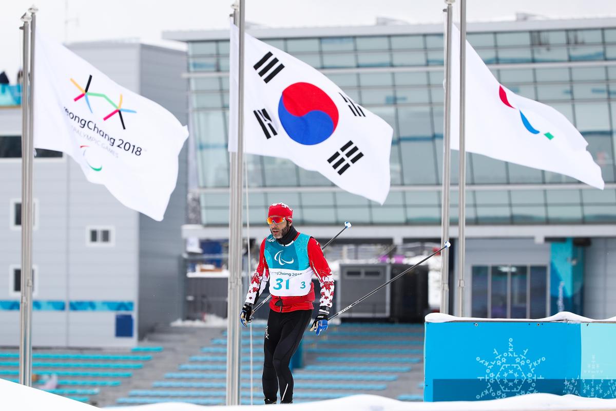 a male Para Nordic skier walks past the Paralympic and South Korean flags