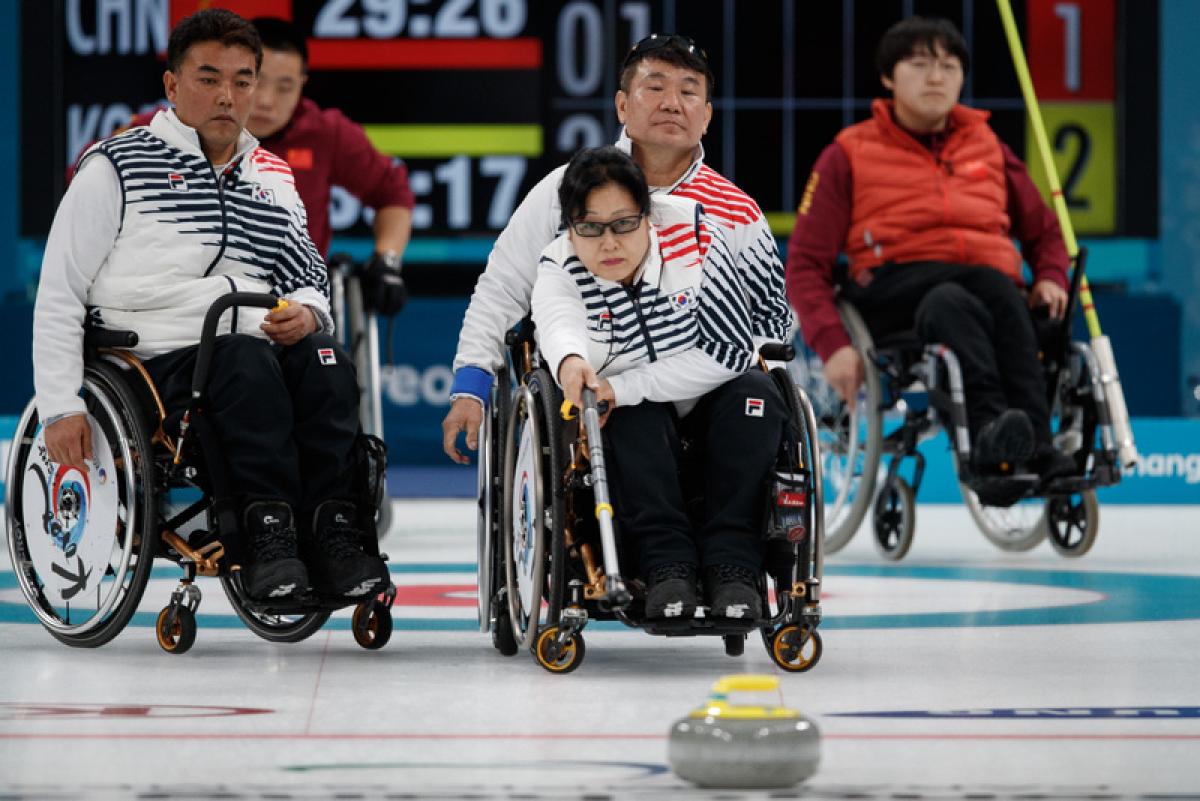 Woman in wheelchair playing curling with three people in wheelchairs observing