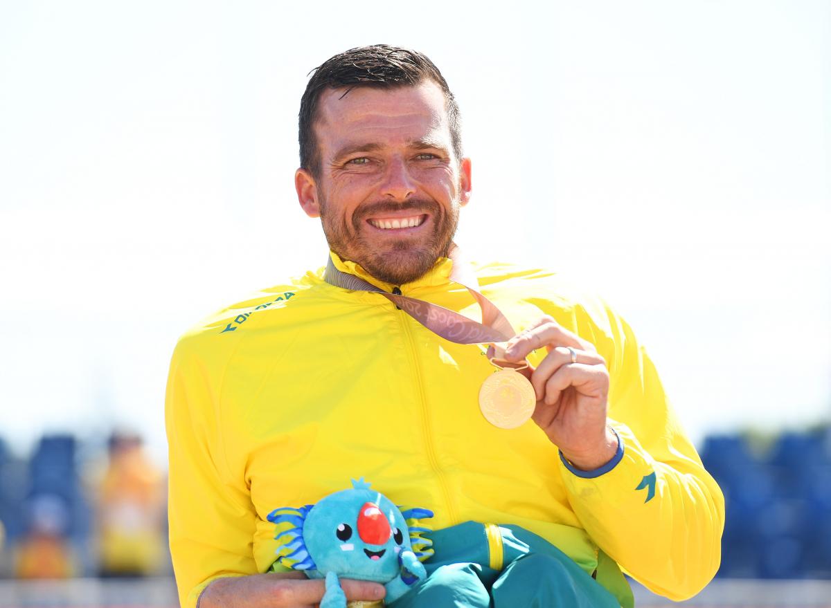 a male wheelchair racer holds up a gold medal