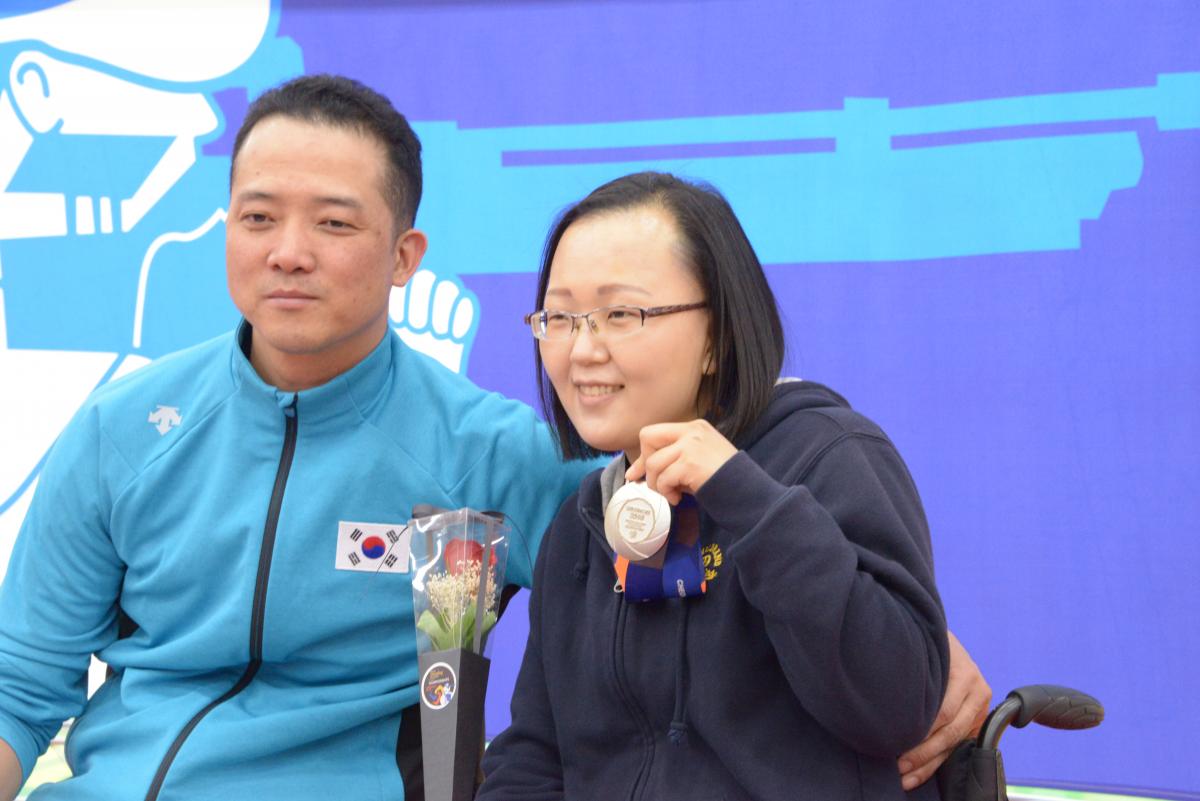 Man and woman in wheelchairs pose for photo with silver medal