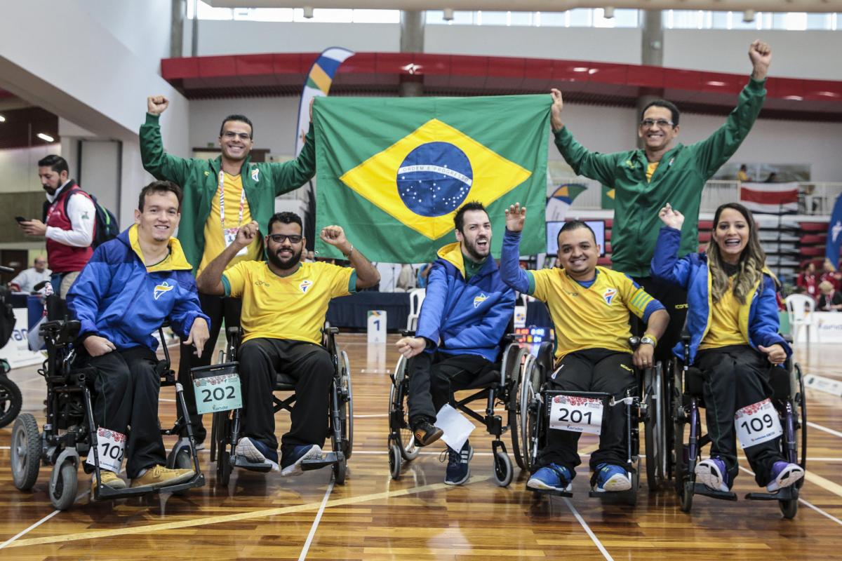 a group of boccia players wave their arms and hold up a Brazil flag