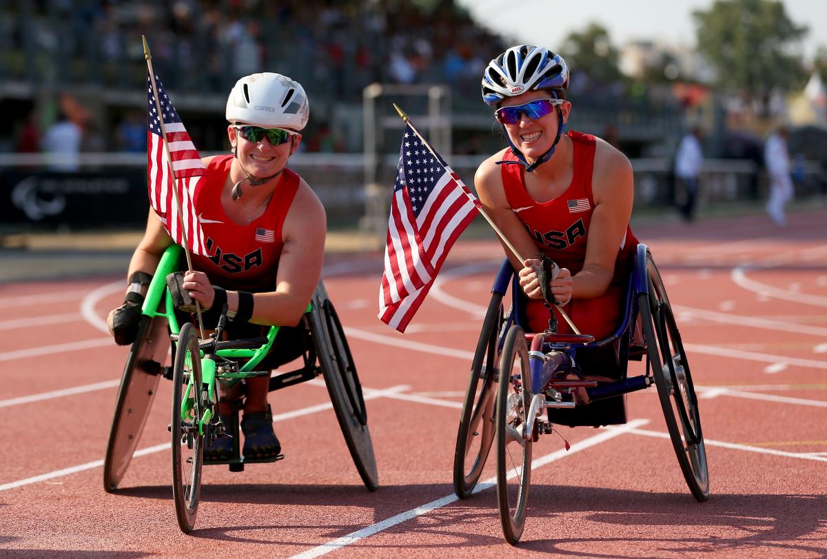 two female wheelchair racers hold up American flags on the track