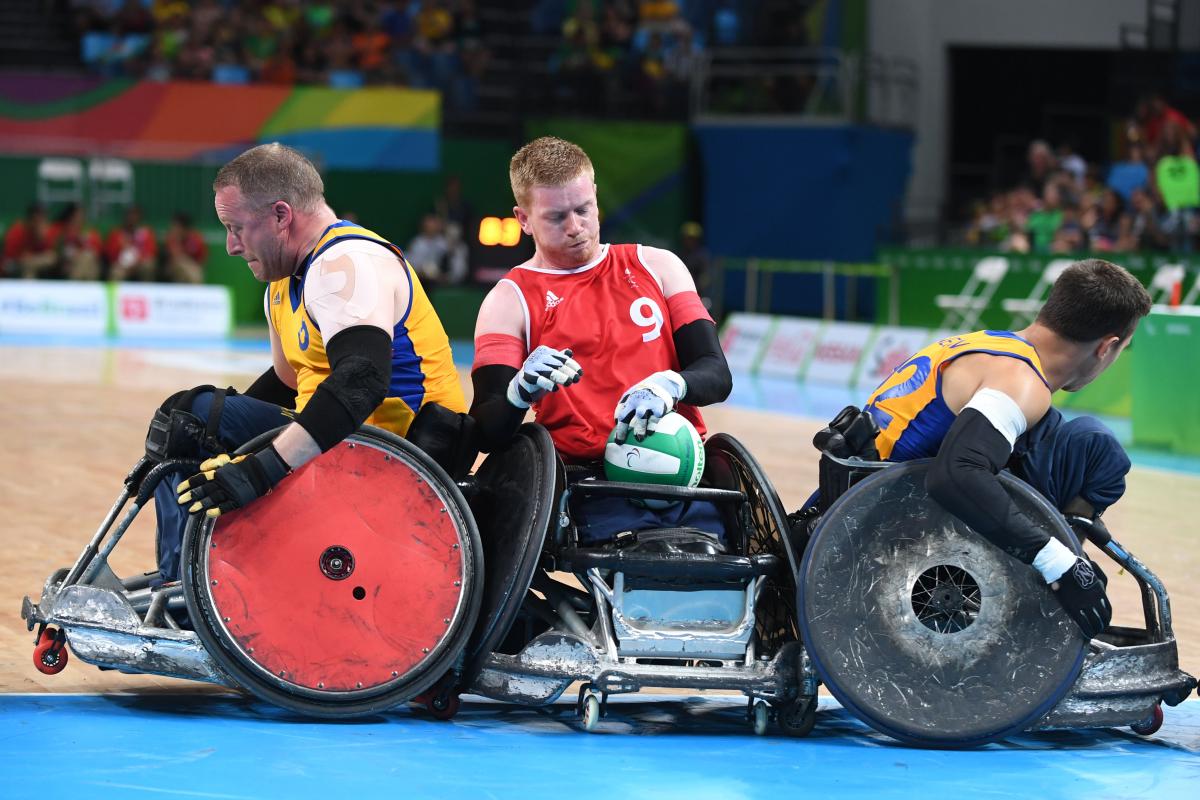 a male wheelchair rugby player holds onto the ball while being challenged by two other players