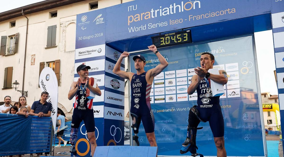 three male Para triathles on a podium with the middle one raising his arms