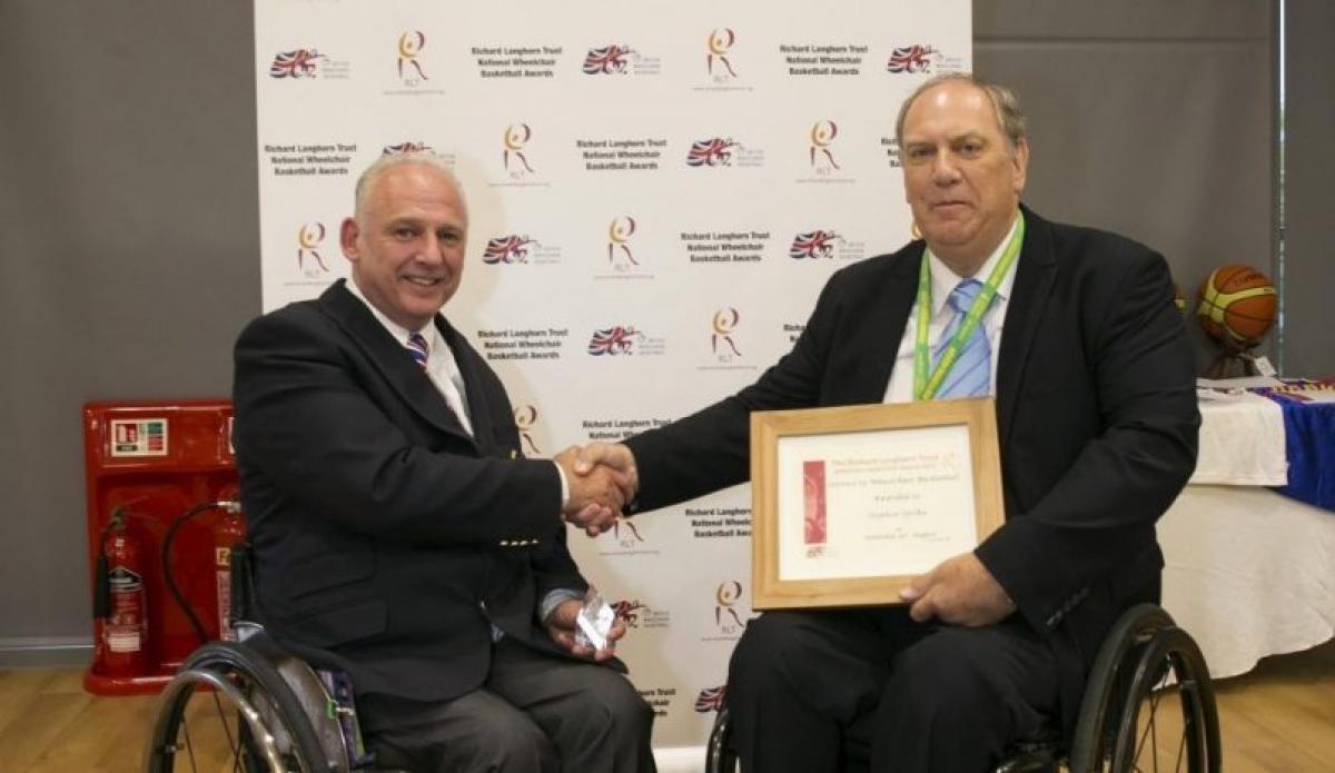 Two men in wheelchair shake hands as one holds a frame