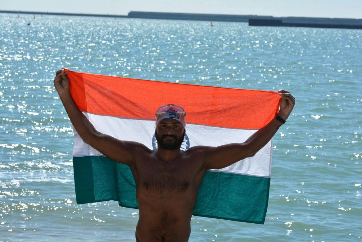 a male Para swimmer holds up in India flag beside the sea