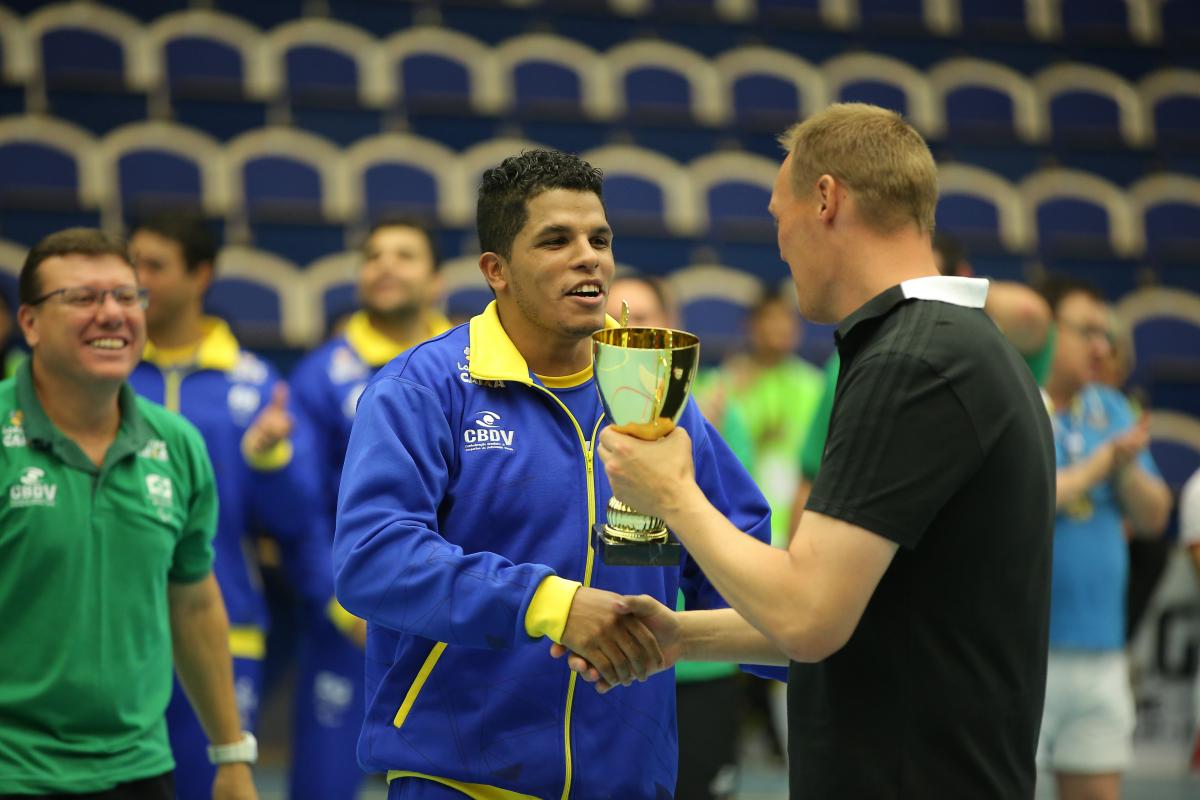 male goalballer player Leomon Moreno receives a trophy and shakes hands with another man