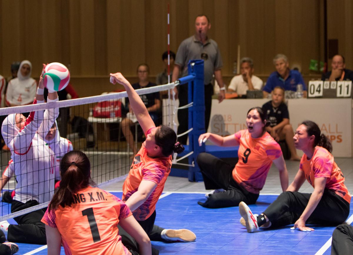 a female Chinese sitting volleyball player punches the ball over the net as her teammates watch
