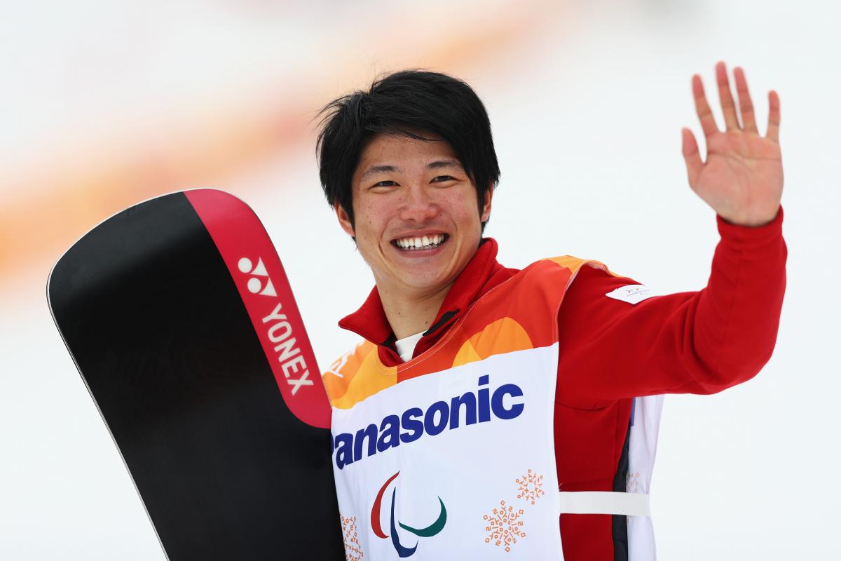 male Para snowboarder Gurimu Narita holds his snowboard and waves to fans