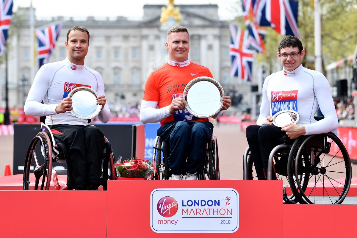 three male wheelchair racers including Marcel Hug and David Weir on a podium holding trophies