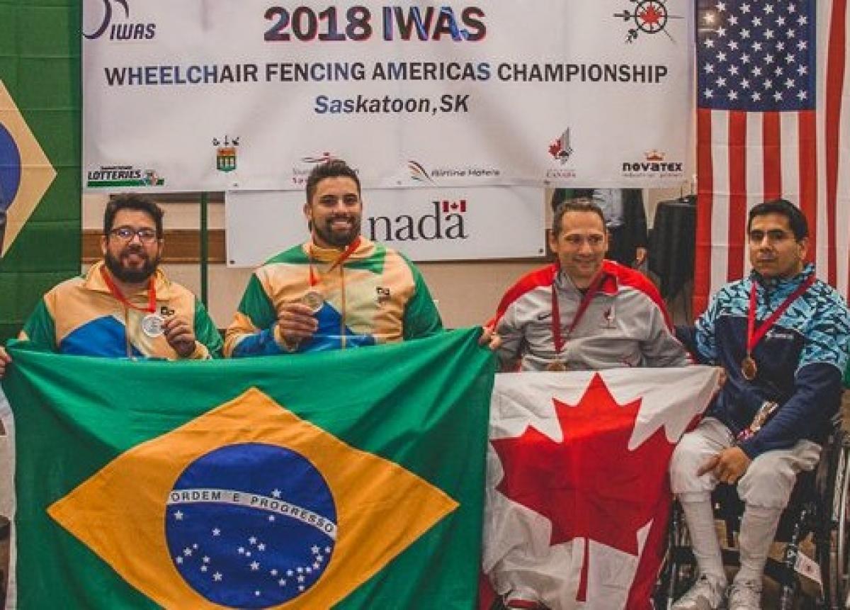 Jovane Guissone - Saskatoon 2018 four male wheelchair fencers on the podium including Jovane Guissone holding a Brazilian flag