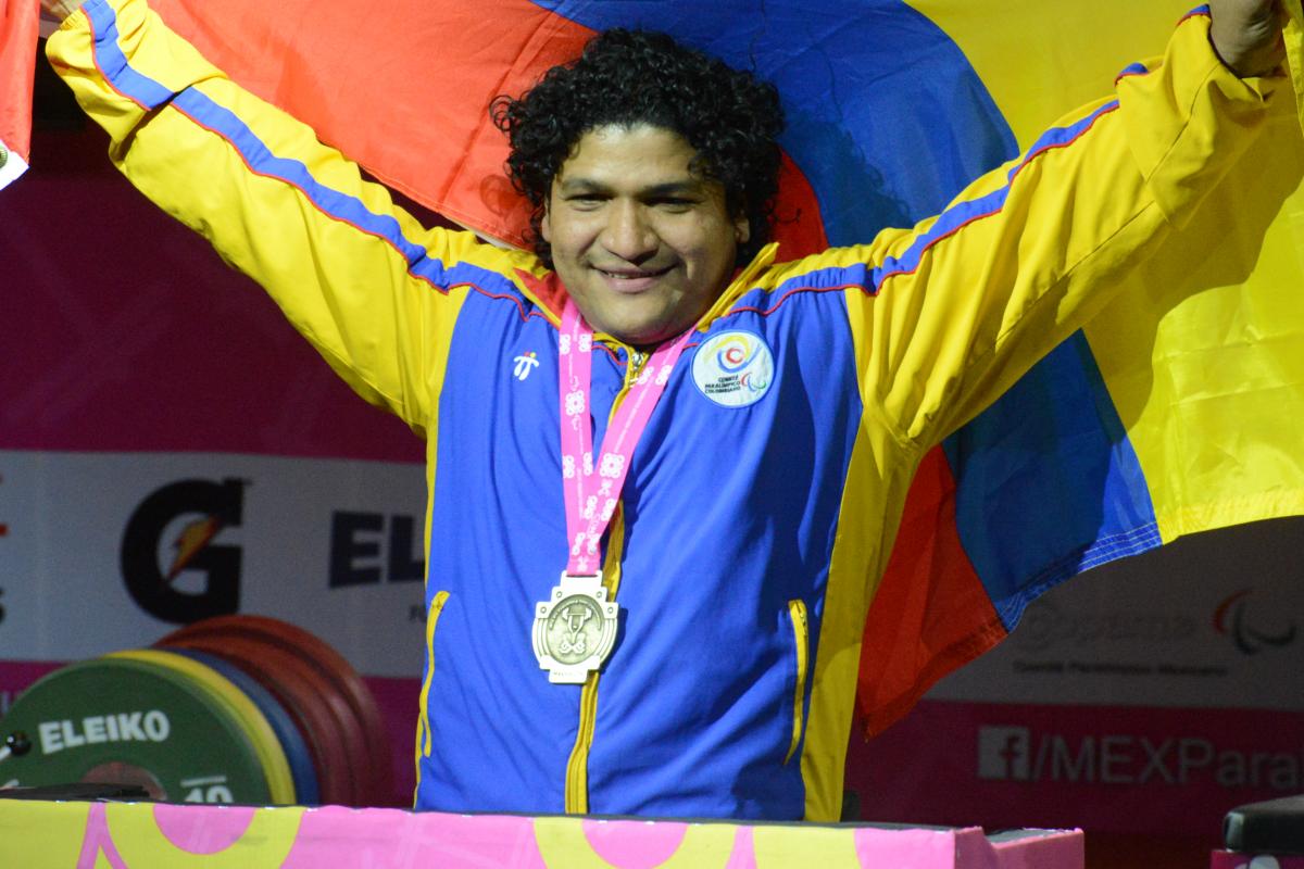 a male powerlifter smiling with his medal round his neck holding up a Colombian flag