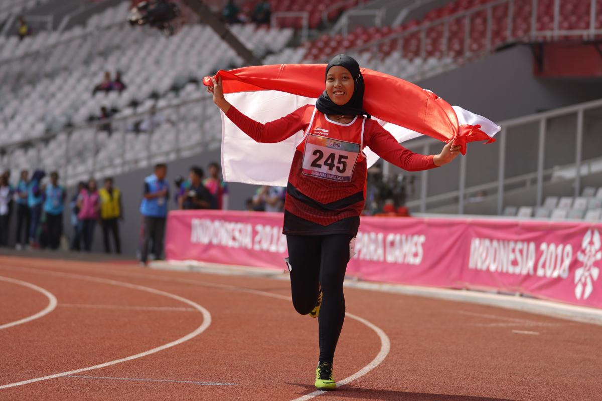 female Para athlete Karisma Tiarani runs down the track holding an Indonesia flag