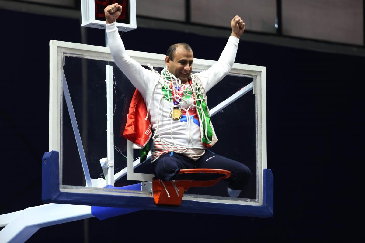 male wheelchair basketball player Morteza Ebrahimi sitting on top of the basketball hoop raising his arms