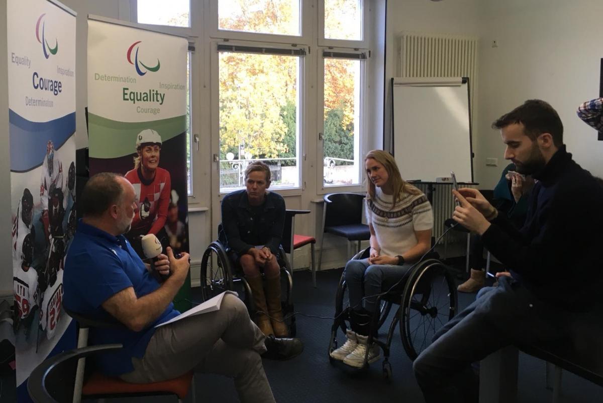 two athletes in wheelchairs watching as a man with a microphone is filmed on a phone