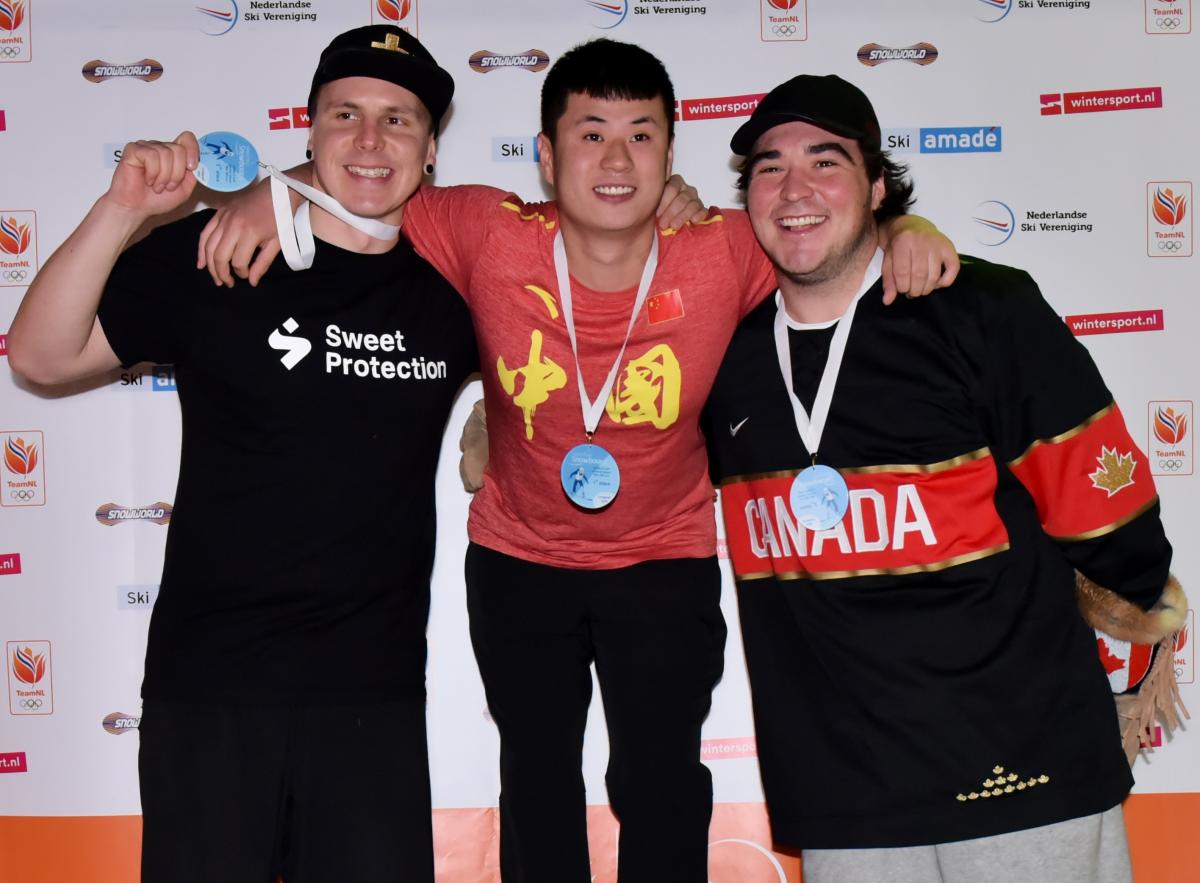 male Para snowboarder Sun Qi stands on the top of the podium with his arms around two other snowboarders