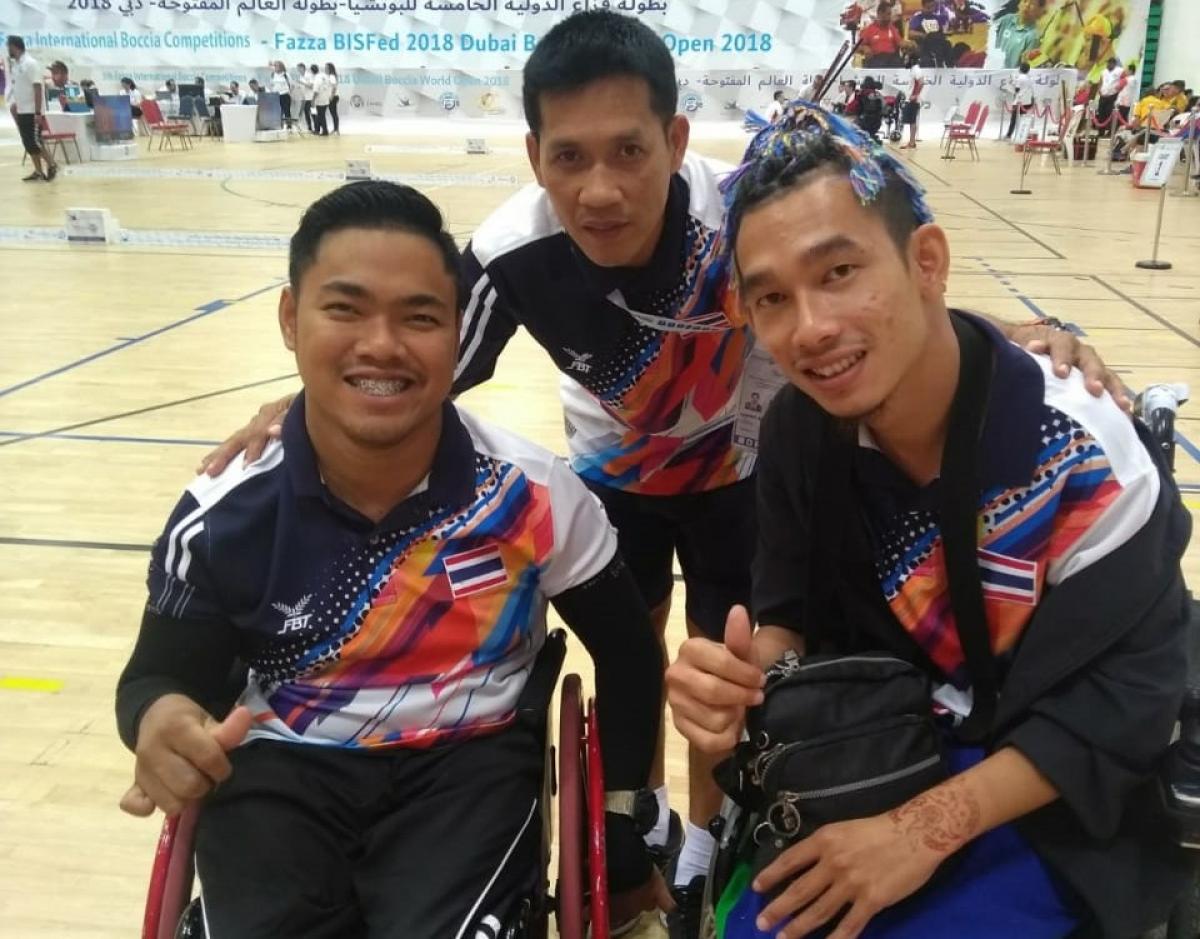 Thailand boccia team two Thai boccia players and their coach standing behind them and smiling with his arms around their shoulders