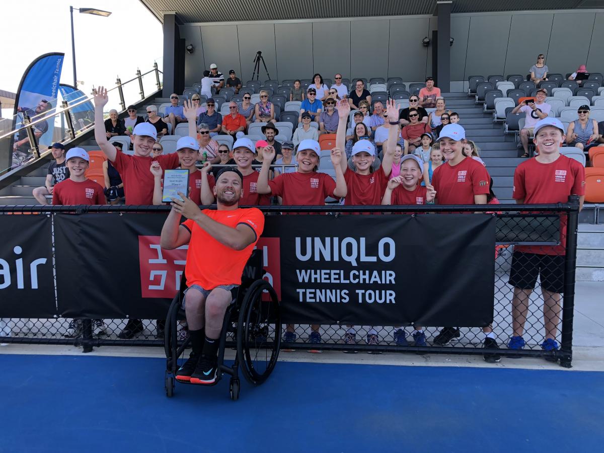 male wheelchair tennis player Dylan Alcott holds up a glass trophy in front of a group of schoolchildren