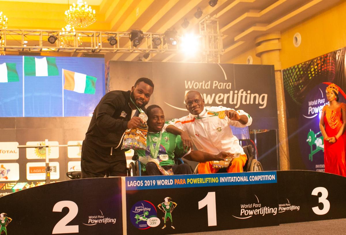 three male powerlifters, with Mufutau Rasaq in the middles, link arms on the podium