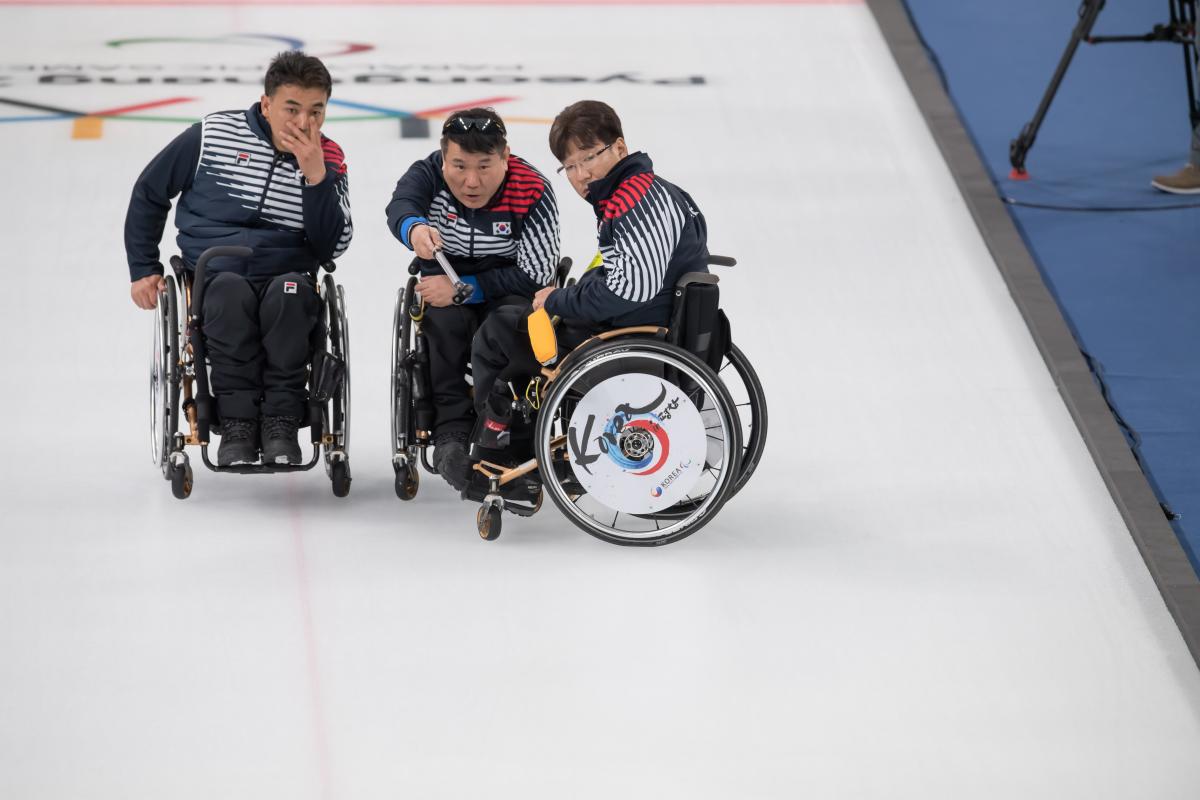 three male wheelchair curlers with Soon-Seok Seo on the right discussing how to play a stone