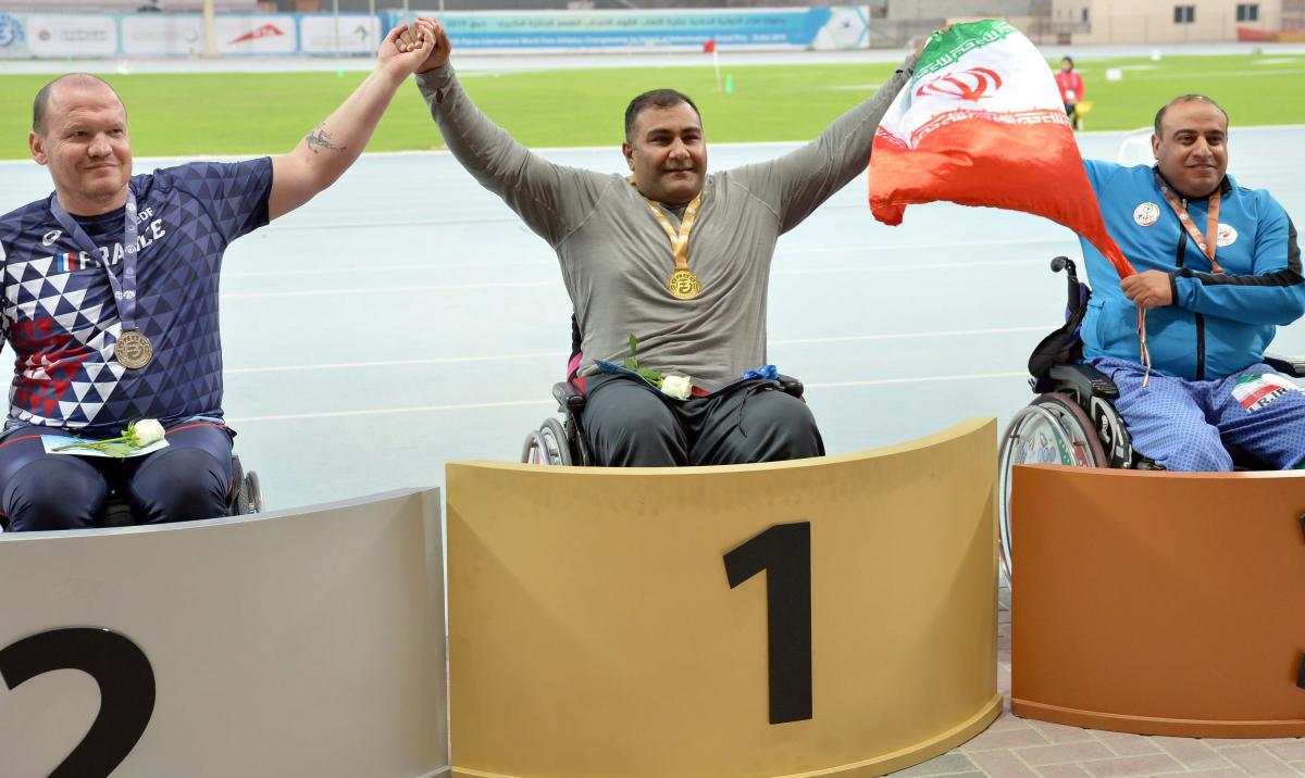 three male Para athletes in wheelchairs on the podium with Hamed Amiri in the centre holding an Iranian flag