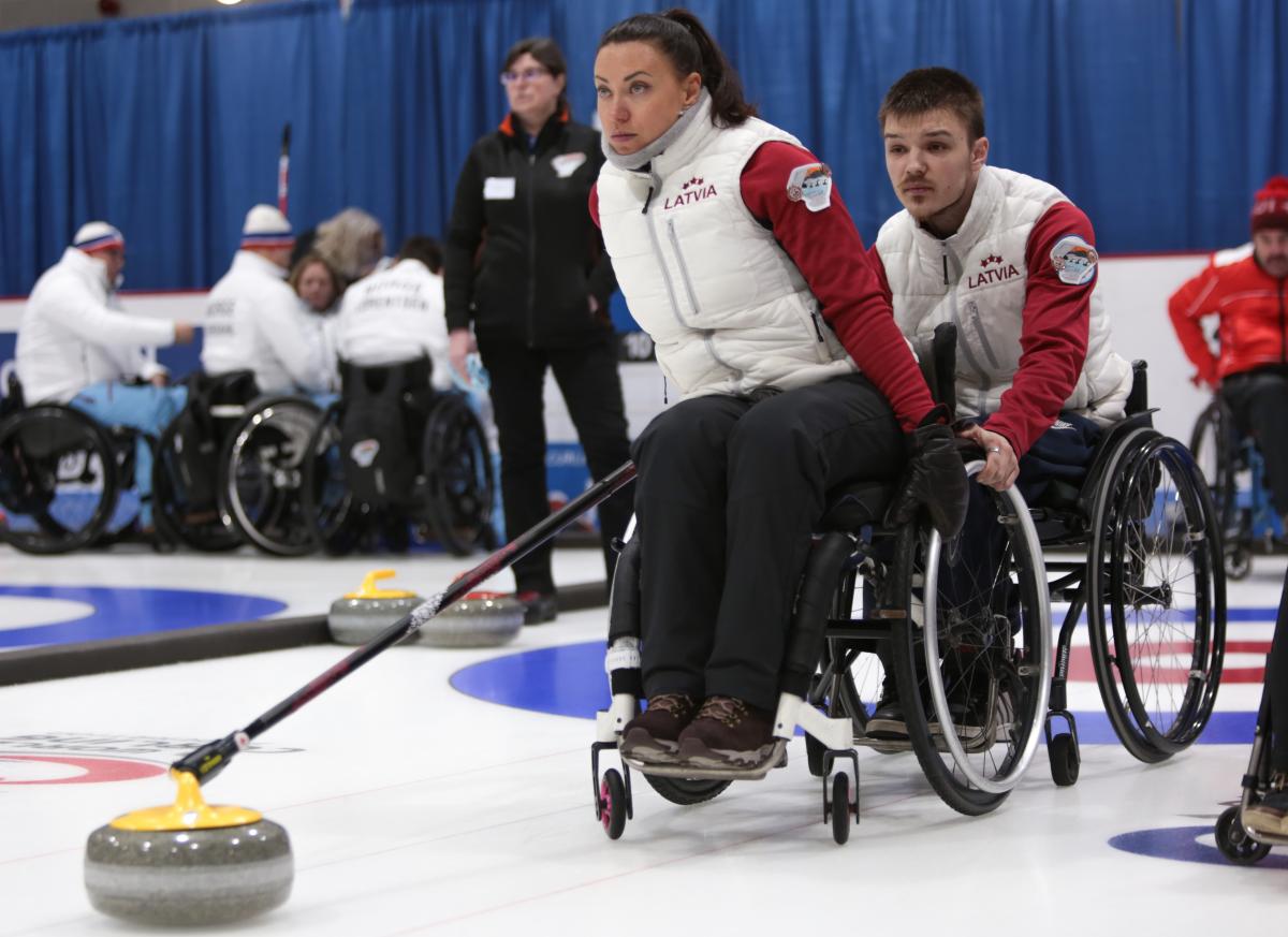 a female wheelchair curler from Latvia plays a stone while another curler holds her wheelchair to steady it
