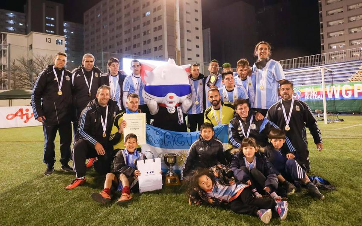 Argentina - 2019 World Grand Prix A group of male blind footballers stand with a blindfolded mascot holding a trophy and the Argentinian flag