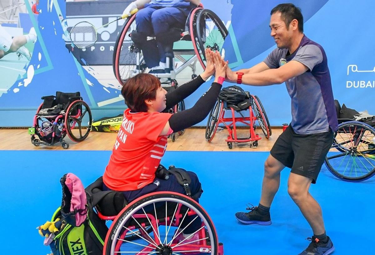female Para badminton player Yuma Yamazaki sits in a wheelchair and high fives a standing man