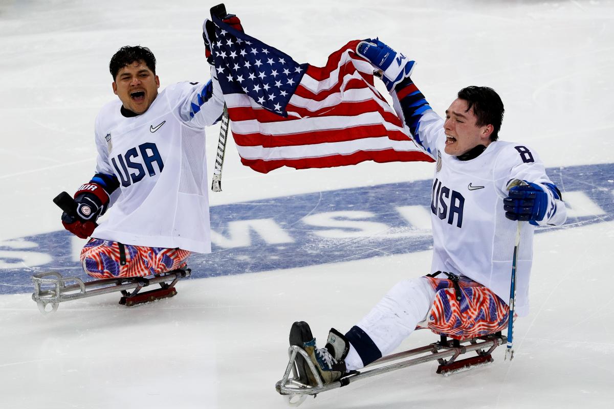 two male US Para ice hockey players on the ice holding a USA flag