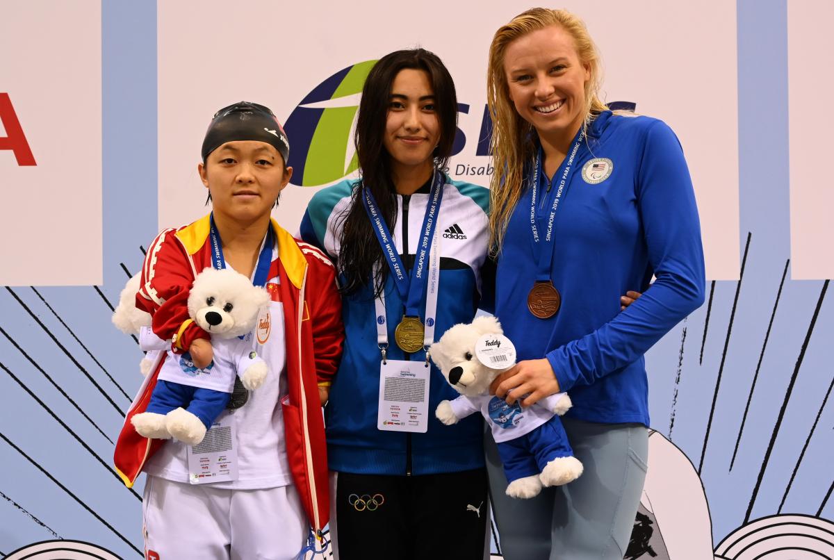 three female swimmers on the podium holding their medals and mascot teddys