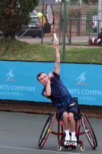 A picture of a man in a wheelchair serving during a wheelchair tennis game