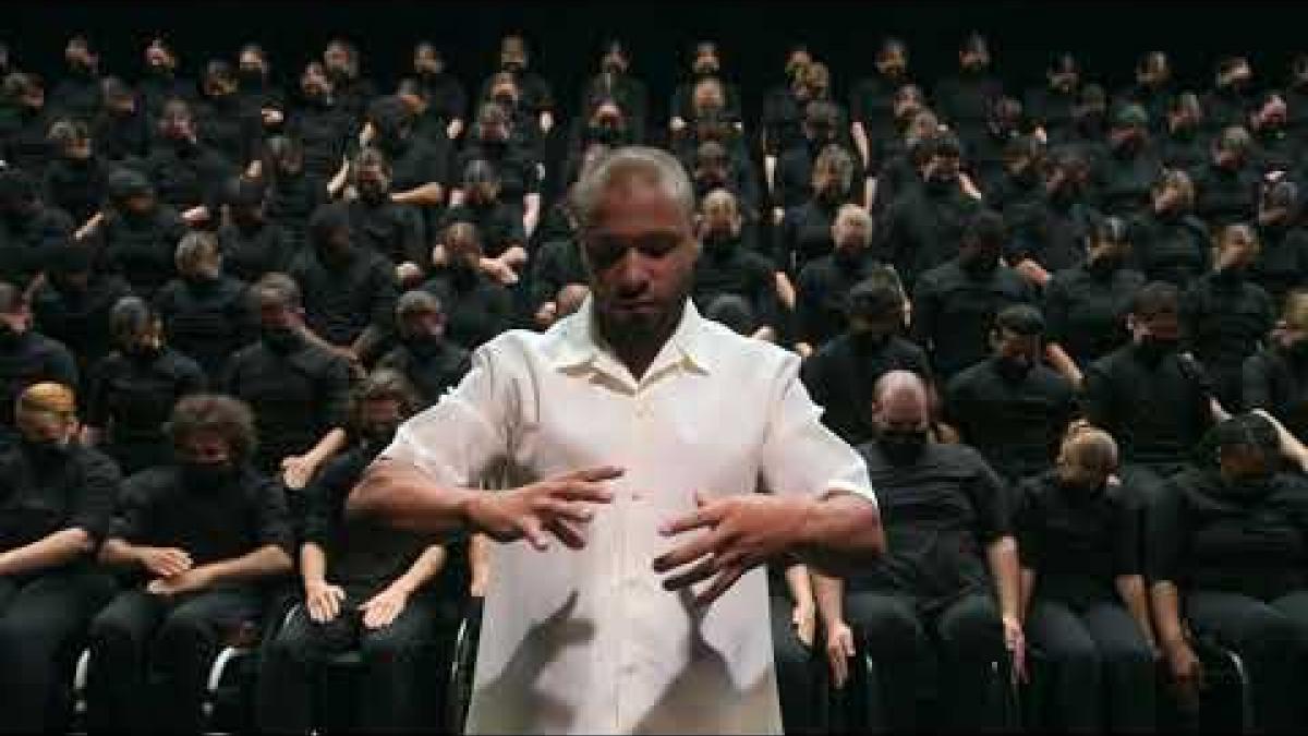 Sadeck Waff, in white, stands with his back to seven rows made up of a total of 128 dancers, the front row made up of dancers in wheelchairs, as he directs them through a dance featuring geometric, synchronised hand movements. 